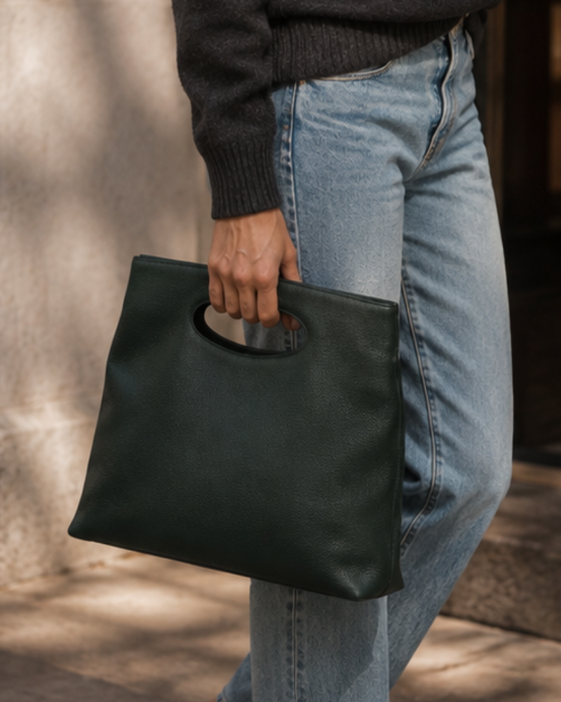 A person wearing a dark gray sweater and light blue jeans holds a dark green pebbled leather tote bag with a circular cutout handle.